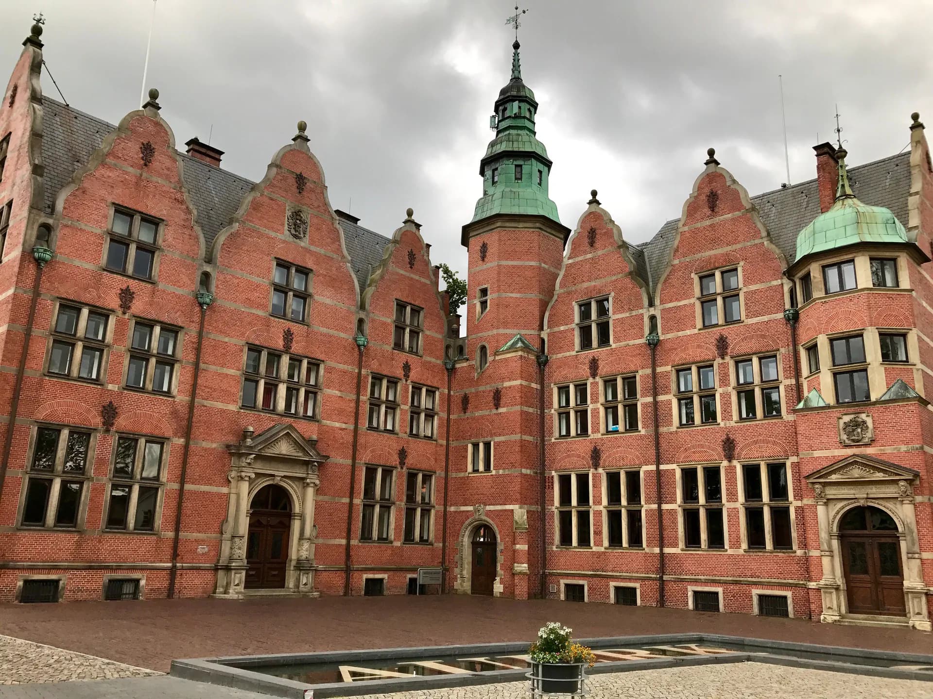 Backstein-Gebäude mit Turm und grünen Kupferkuppeln, vor bewölktem Himmel.